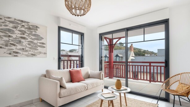 Un salon moderne comprenant un canapé, une petite table, un fauteuil en osier et une grande fenêtre donnant sur un balcon avec des balustrades rouges.