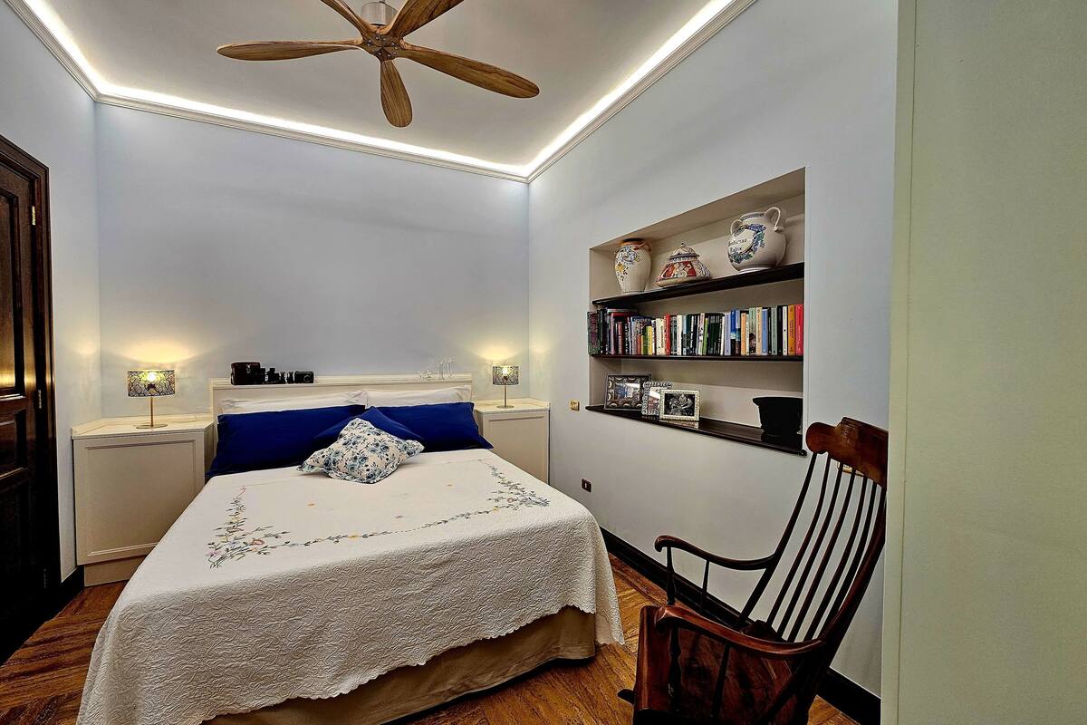 Un dormitorio con cama, mesitas de noche, estantería, una silla y ventilador de techo.