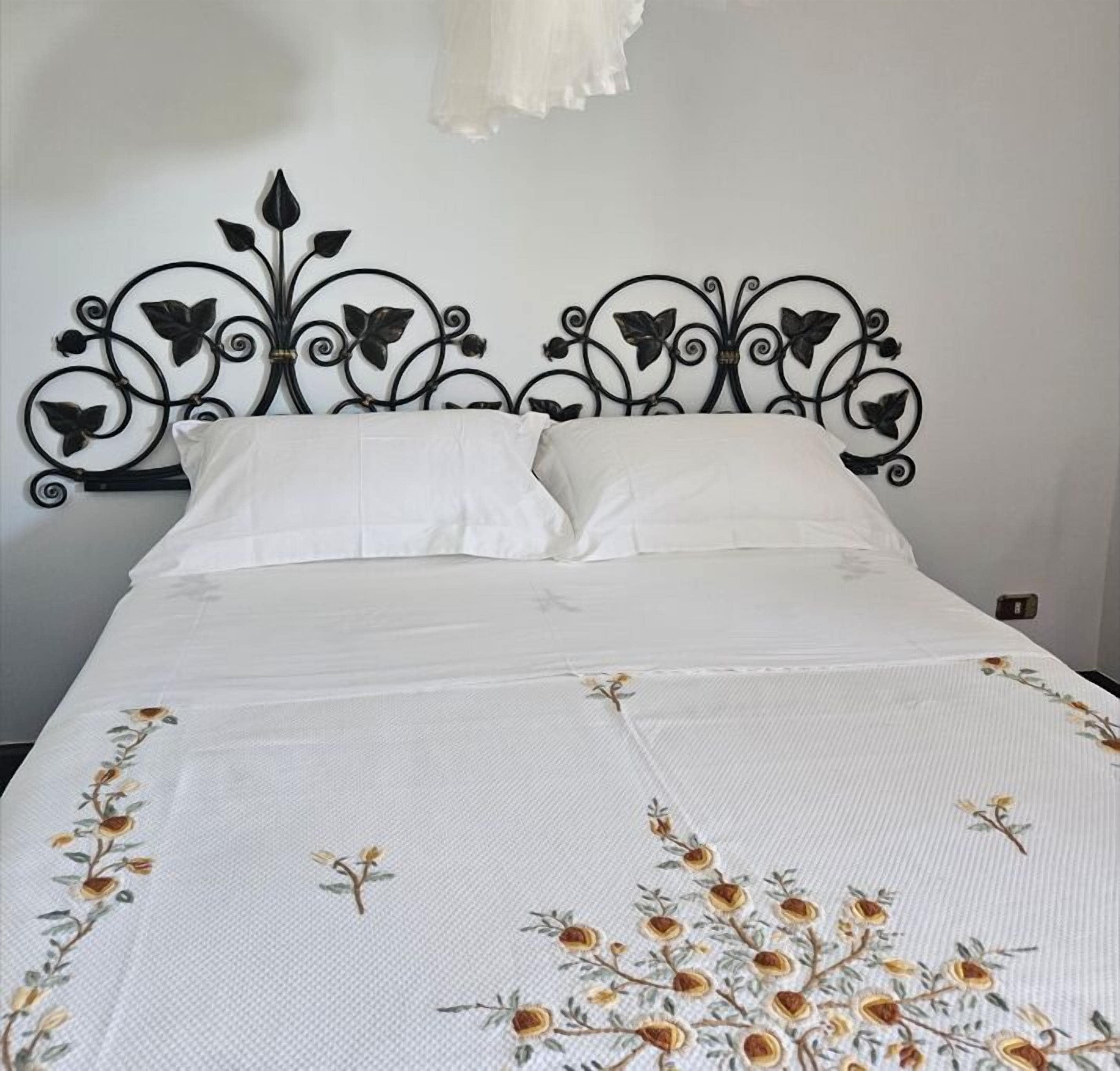 Una cama con colcha blanca con bordados florales, cabecera metálica negra con diseños de hojas y almohadas blancas.