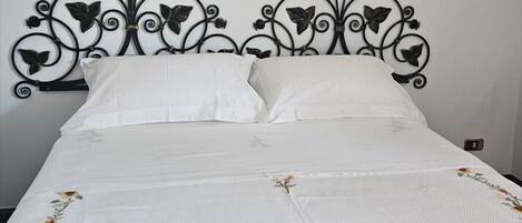 Una cama con colcha blanca con bordados florales, cabecera metálica negra con diseños de hojas y almohadas blancas.