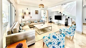 A spacious living room with a sofa set, a coffee table, and a TV. There is a staircase with glass railings leading to an upper floor. - Villa Neptune Pour 8 Pieds Dans l eau Carthage (Carthage)