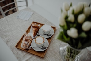 A tray with croissants and coffee cups on a table, accompanied by a vase of white flowers.