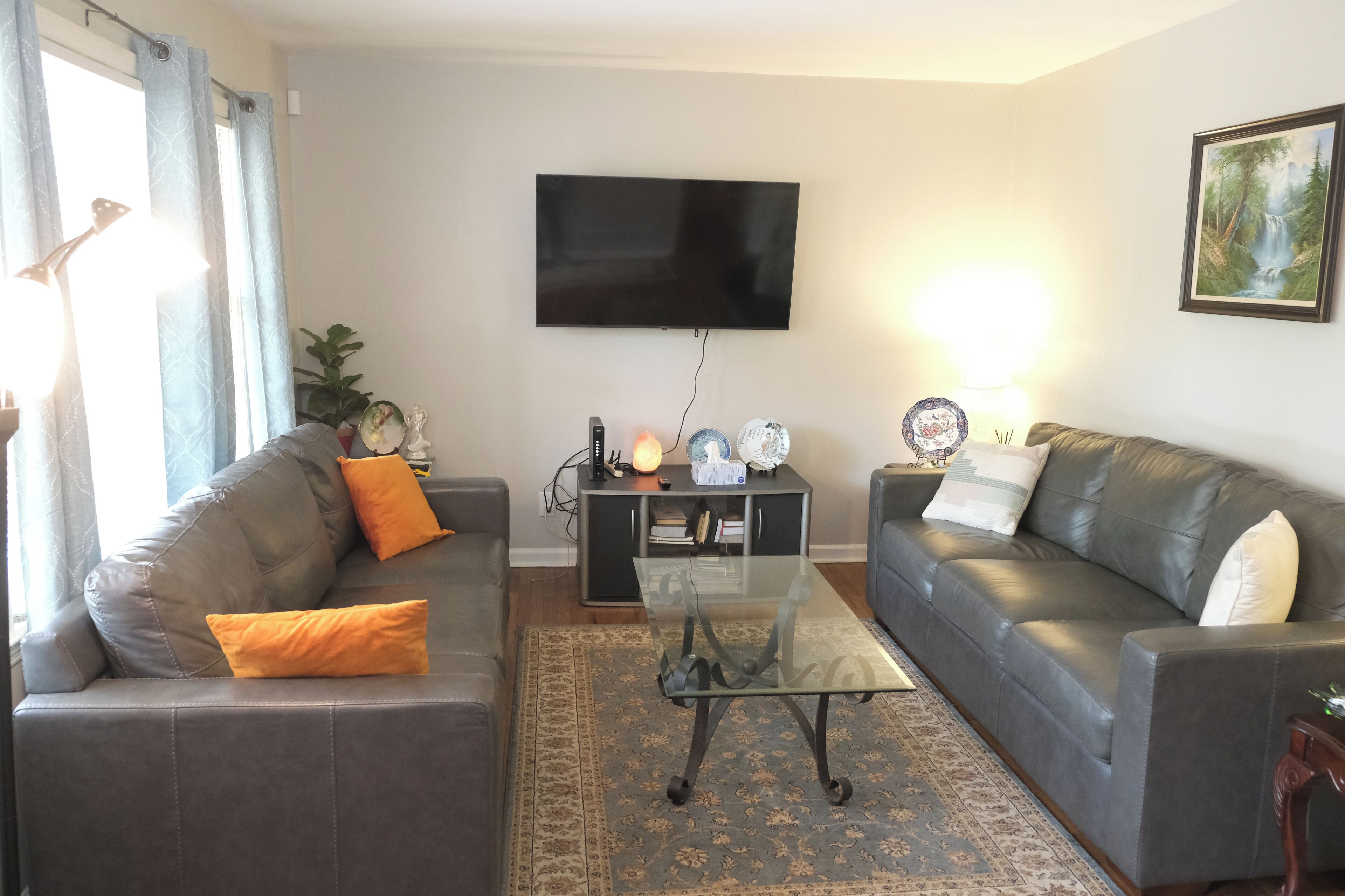 Ein Wohnzimmer mit zwei grauen Sofas, einem Glastisch, einem flachbildfernseher an der Wand, einer kleinen Kommode mit Dekoration und einem gemusterten Teppich.