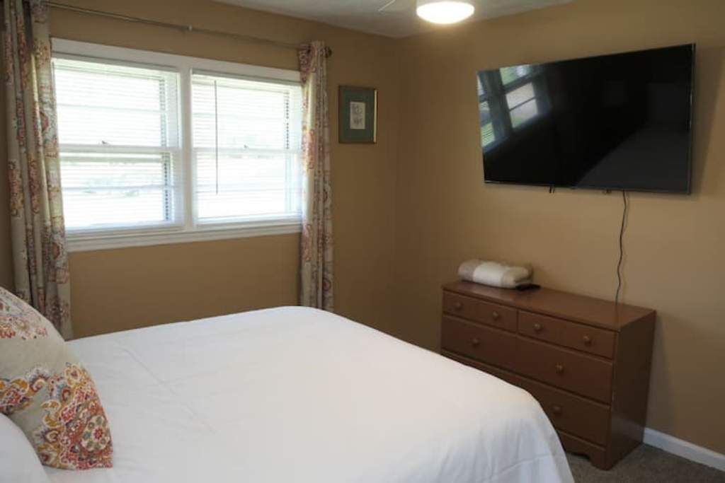 A bedroom with a bed, a dresser, a television mounted on the wall, and a window with curtains.