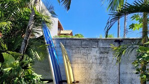 A concrete wall with a surfboard leaning against it, a coiled garden hose, and a palm tree in the foreground. - Pousada Makani Kai (Florianópolis)