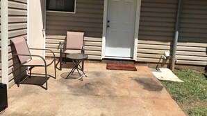 Une terrasse couverte avec deux chaises et une petite table, une porte blanche et une fenêtre avec des stores.