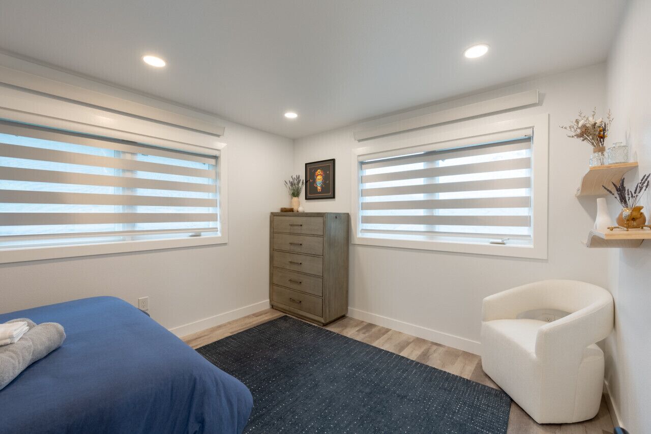 A bedroom with a bed, a dresser, a chair, and a window with blinds.