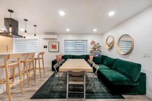 Un soggiorno moderno con tavolo da pranzo in legno, divano verde e angolo cucina con mobili in legno e lampade a sospensione.