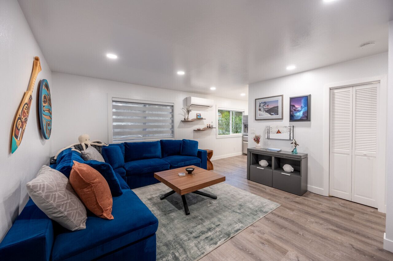 A modern living room with a blue sofa, a wooden coffee table, and a grey rug. There is a grey cabinet with decorative items and a window with blinds.