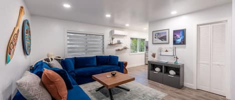 Een moderne woonkamer met een blauwe bank, een houten salontafel en een grijze vloerkleed. Er staat een grijs kastje met decoratieve objecten en een raam met jaloezieën.