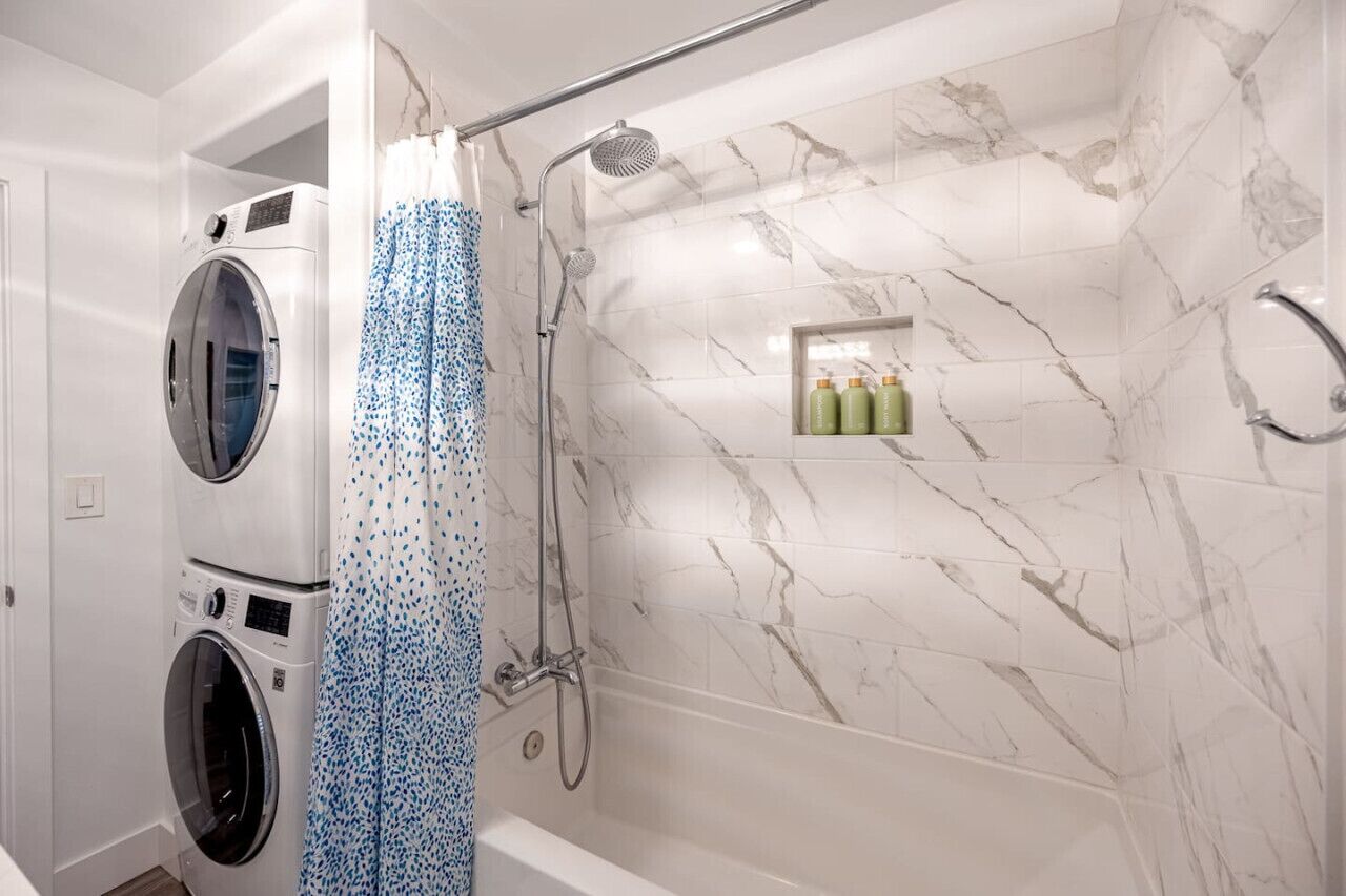 A bathroom with a shower, two stacked washing machines, and a marble-tiled wall.