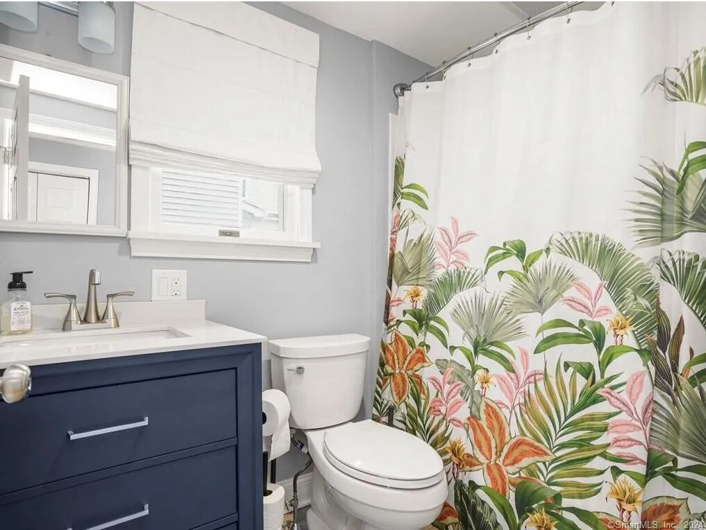 Une salle de bain avec des toilettes blanches, un meuble-lavabo bleu foncé doté de tiroirs, un lavabo blanc et un rideau de douche orné d’un motif de feuilles tropicales.