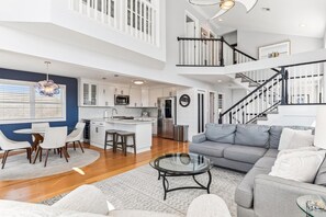 A spacious living area with a grey sofa, a glass coffee table, and a dining area with a round table and chairs. A kitchen with white cabinets and a stainless steel refrigerator is visible in the background. - Bayside Retreat in Bethany Beach: Short Walk to Beach, Sleeps 13, Water Views! (Bethany Beach)
