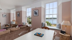 A cozy living room with a fireplace, a sofa, a dining table, and large windows overlooking a scenic view. - Laston Garden Apartment - Tenby Harbour, Sea Views (Tenby)