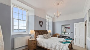 A bedroom with a large window, a bed, a nightstand, a dresser, and a mirror. - Laston Garden Apartment - Tenby Harbour, Sea Views (Tenby)
