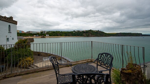 A balcony with a table and chairs overlooking a body of water, a white building, and a distant shoreline. - Laston Garden Apartment - Tenby Harbour, Sea Views (Tenby)