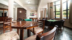 Een ruime eethoek met houten tafels en stoelen, grote ramen met gordijnen en uitzicht op groen.