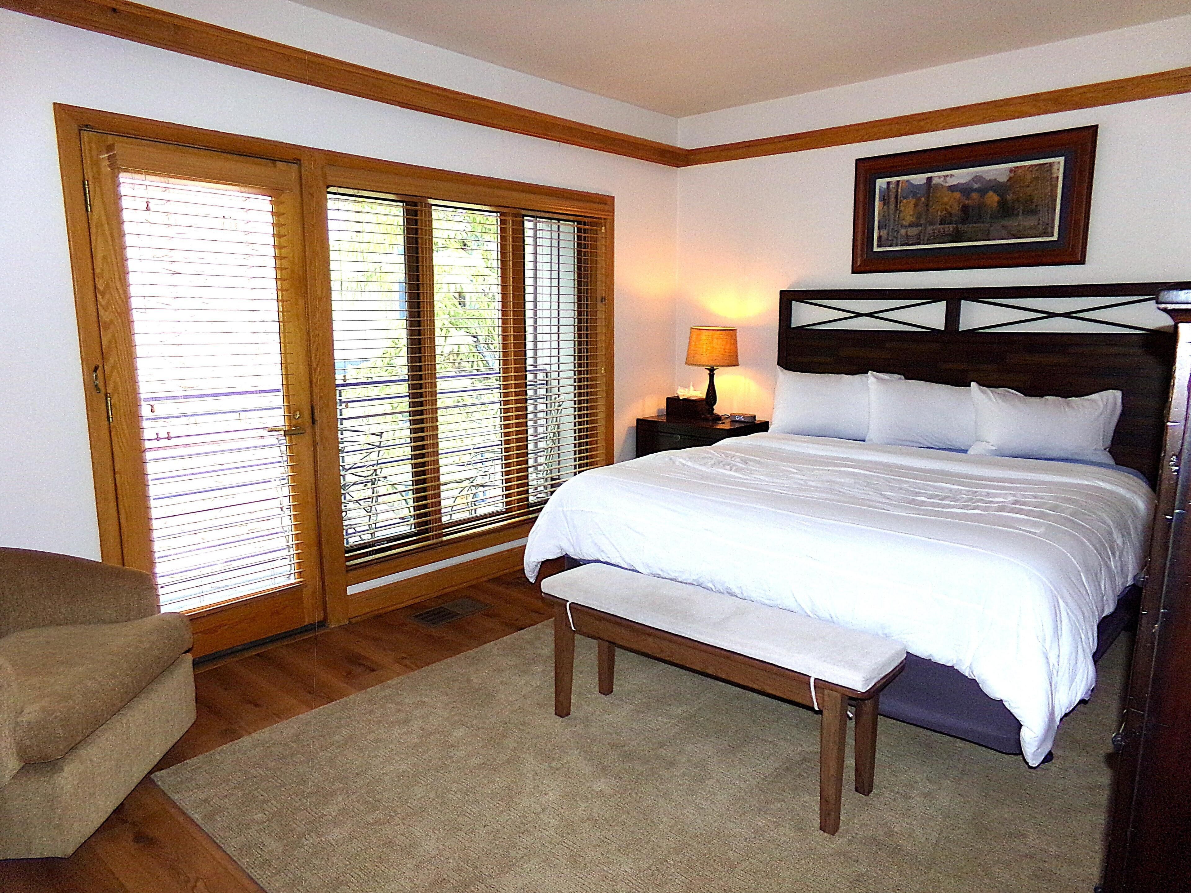 A bedroom with a large bed, a bench, a nightstand with a lamp, and a painting on the wall.