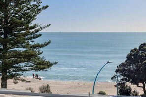 A beach with a clear view of the ocean, a few people walking, and a large tree in the foreground.
