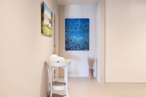 A hallway with a white wicker chair, a painting, and a vase with dried plants.