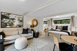 A modern hotel room with a sofa, ottoman, and a bed with a view of greenery outside. - 1-bed wow factor in Kohimarama (Auckland)