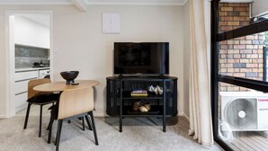 En kompakt opholdsstue med spiseplads, et sort tv-møbel og udsigt til en murstensfacade gennem et vindue.