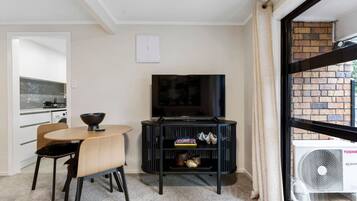 A compact living space with a dining area, a black entertainment unit with a TV, and a view through a window to a brick exterior.