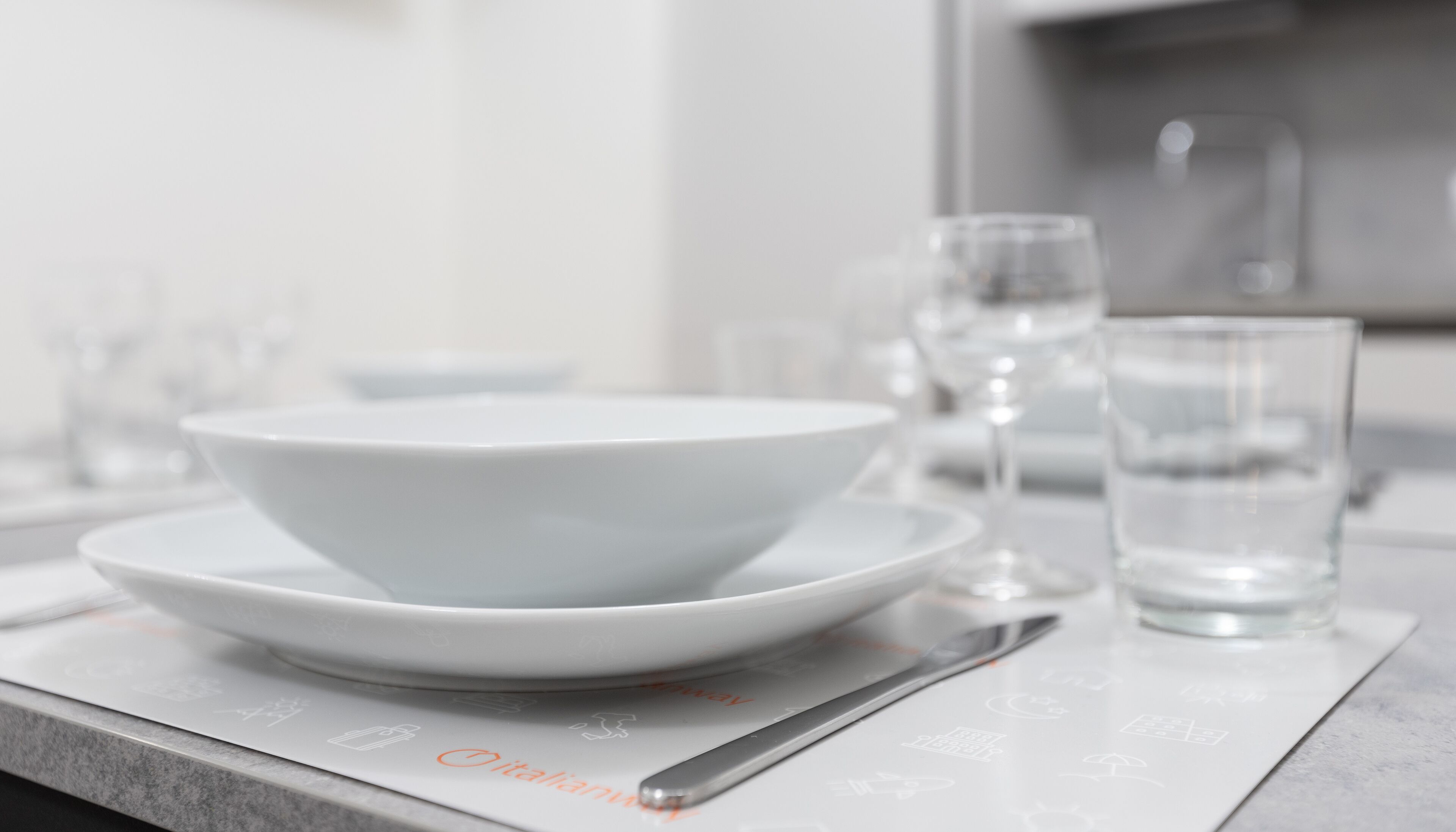 A white bowl on a plate on a table with a spoon and a glass of water.