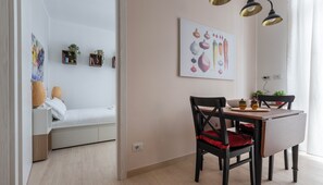 Une pièce moderne et bien éclairée, avec un coin repas comprenant une table et des chaises en bois, un canapé blanc et un tableau représentant des légumes, accroché au mur.
