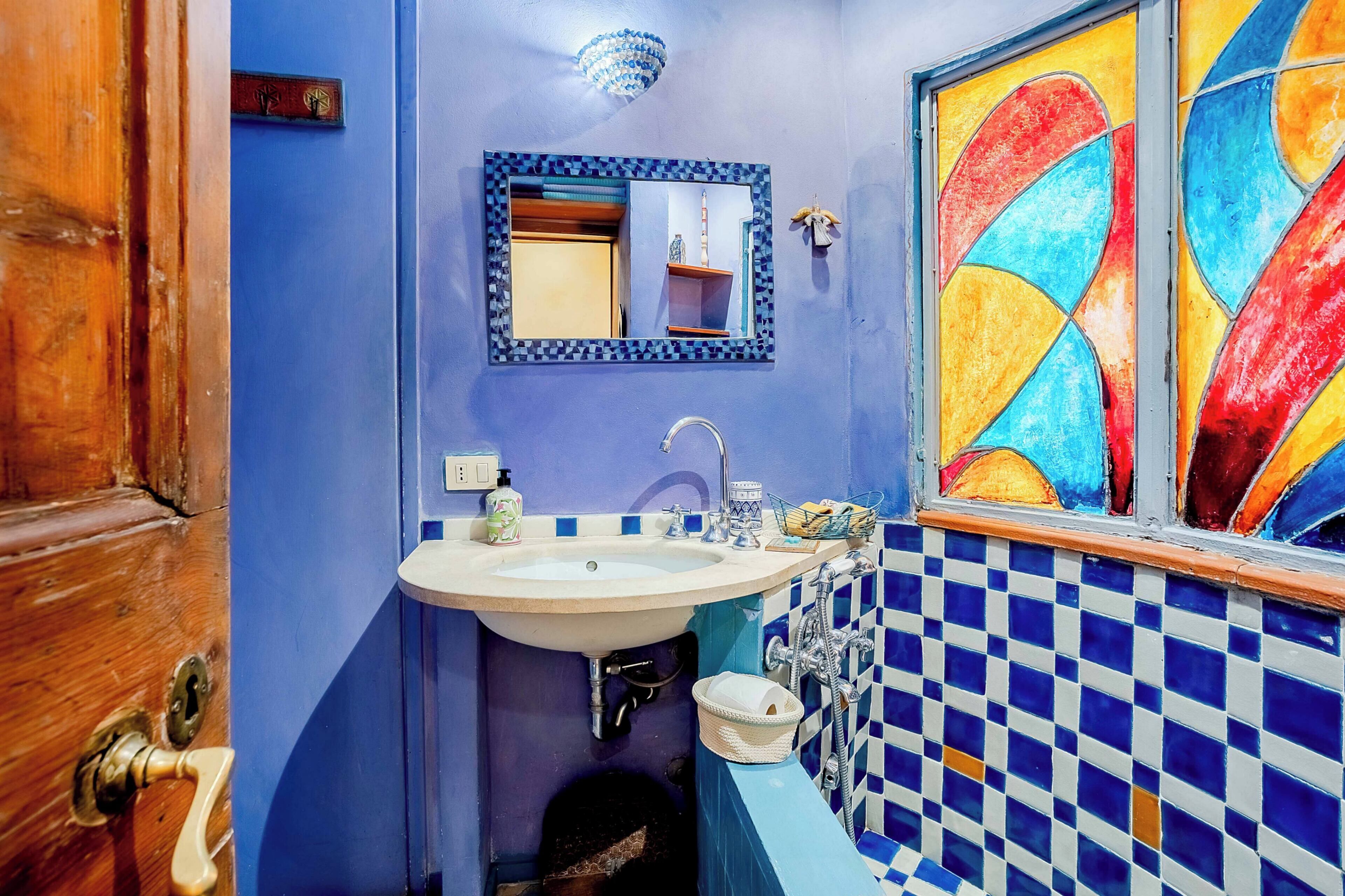 A bathroom with a blue tiled wall, a white sink with a mirror above it, and a colorful window.