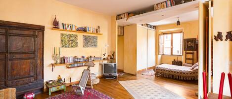 A spacious living room with wooden flooring, a large rug, a television, and a bookshelf filled with books.