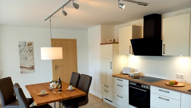 A modern kitchen with white cabinets, a black range hood, and a dining area with a wooden table and chairs. - Ferienwohnung Hansen, in Ruhiger Lage mit Wunderschönem Ausblick (Welschbillig)