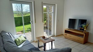 A living room with a grey sofa, a wooden TV stand, and a sliding glass door leading to a garden. - Ferienwohnung Hansen, in Ruhiger Lage mit Wunderschönem Ausblick (Welschbillig)