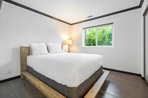 Ein modernes Schlafzimmer mit einem großen Bett, einem Nachttisch, einer Lampe und einem Fenster mit Blick ins Grüne.