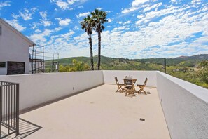 A rooftop terrace with a table and chairs, palm trees, and a view of green hills.