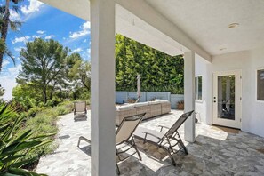 Une terrasse extérieure couverte, dotée d’un sol en pierre, d’un espace salon avec des coussins et d’une porte avec un panneau de verre.