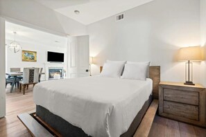 A modern hotel room with a large bed, a bedside table with a lamp, a TV mounted on the wall, and a fireplace.