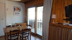 A room with a wooden dining table, chairs, a television, and a window with a view of a wooden deck.