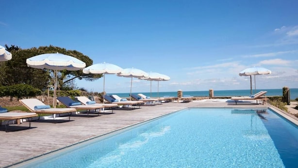 Zona junto a la piscina con sillones de descanso y sombrillas, cielo despejado y vista al mar.