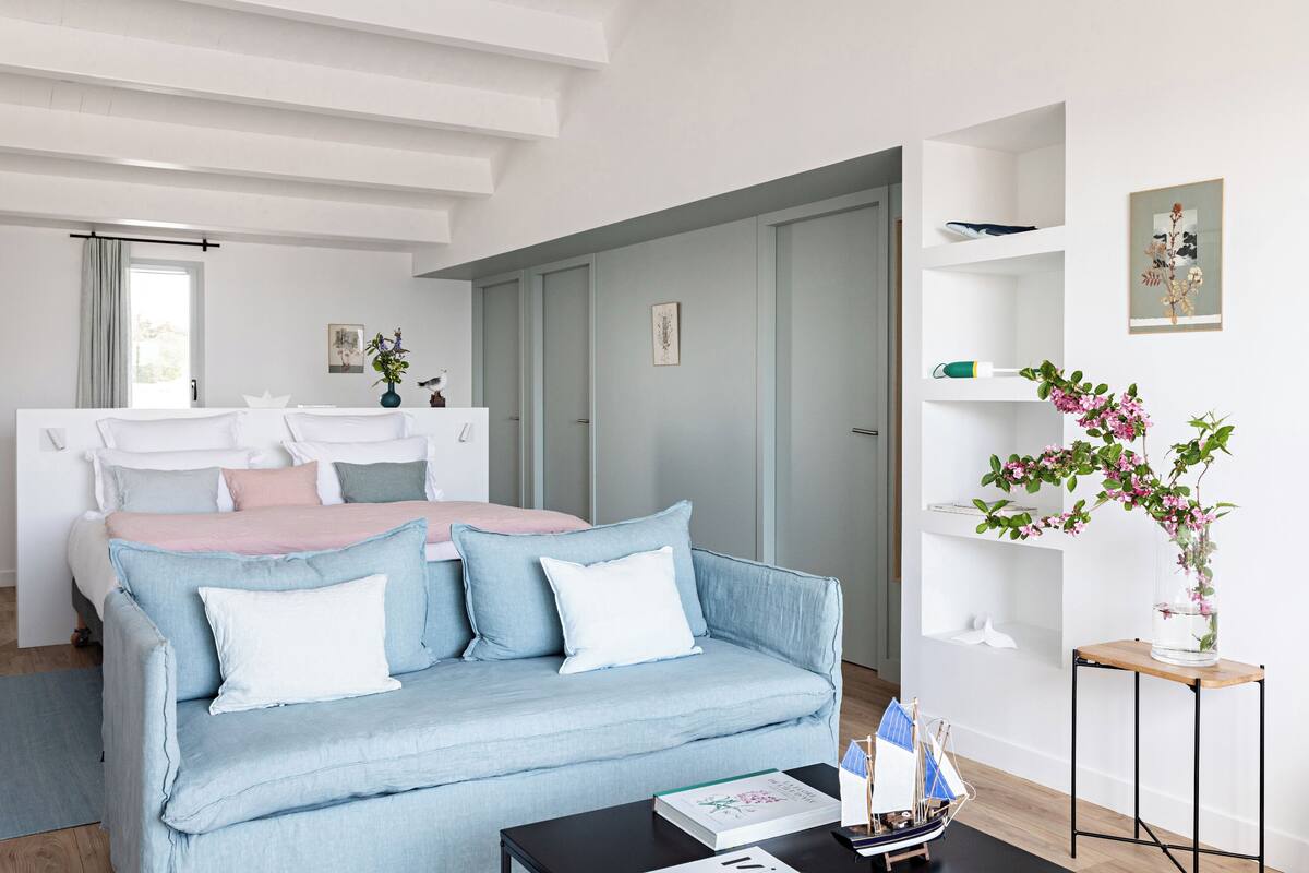A modern bedroom with a light blue sofa, a wooden coffee table, and a bedside table with a book and a ship model.