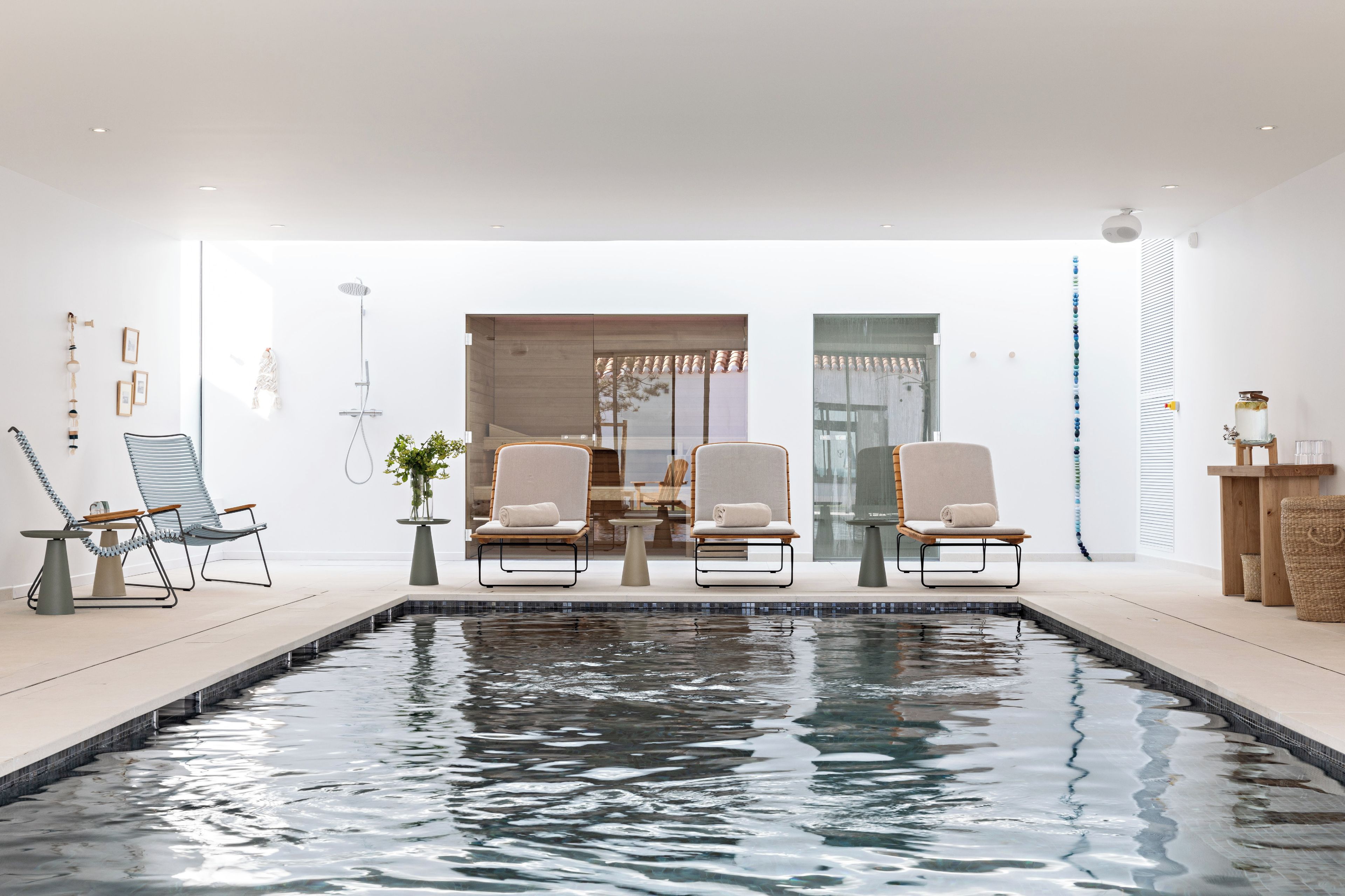 Un espace piscine intérieur moderne, comprenant deux chaises longues, une petite table et un espace douche clos par des parois de verre.