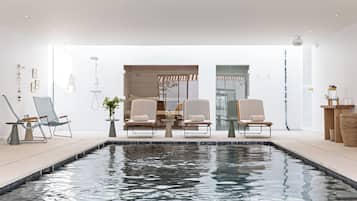 Un espace piscine intérieur moderne, comprenant deux chaises longues, une petite table et un espace douche clos par des parois de verre.