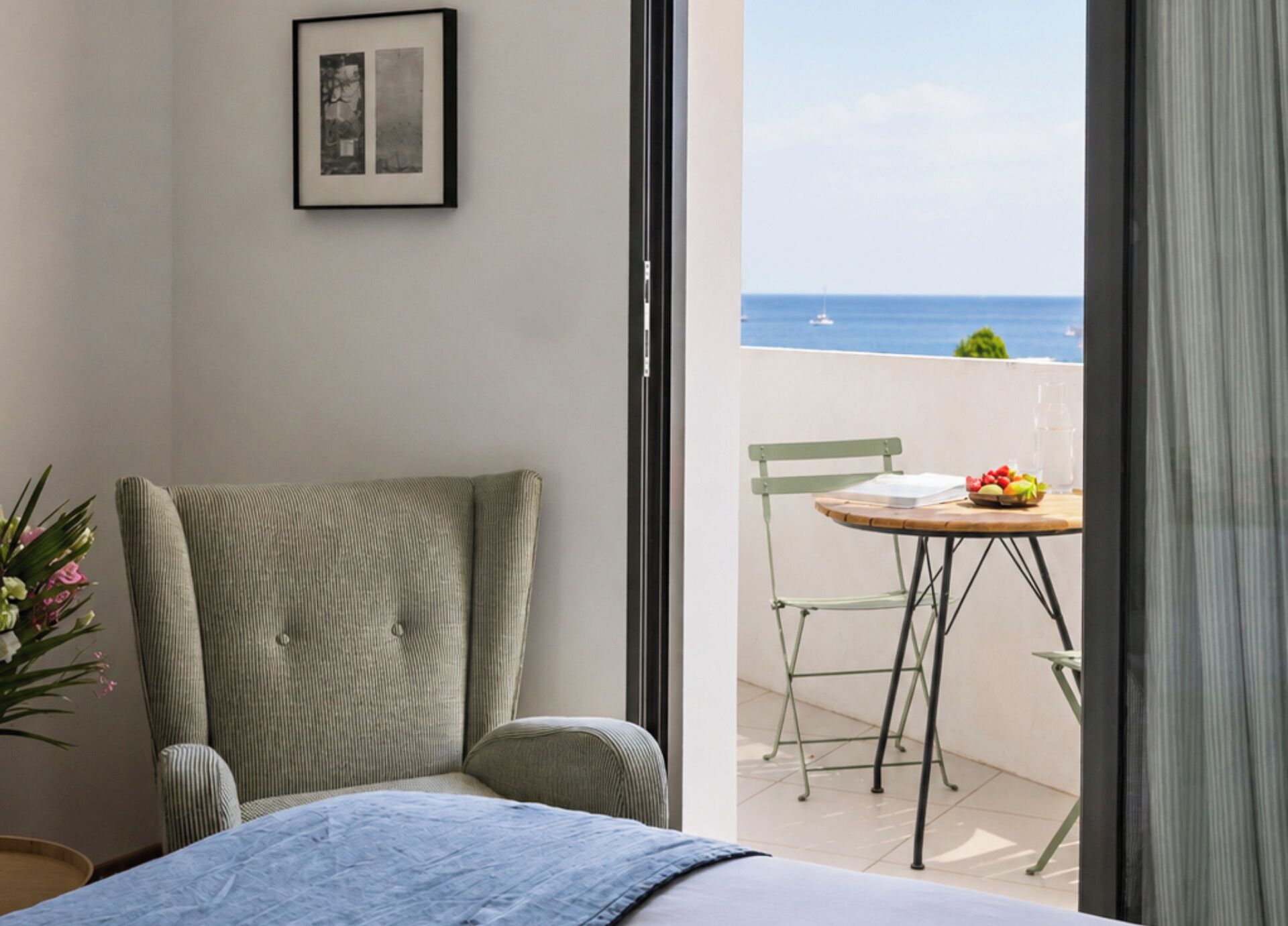 A room with a bed, a chair, a table with fruit, and a view of the sea.