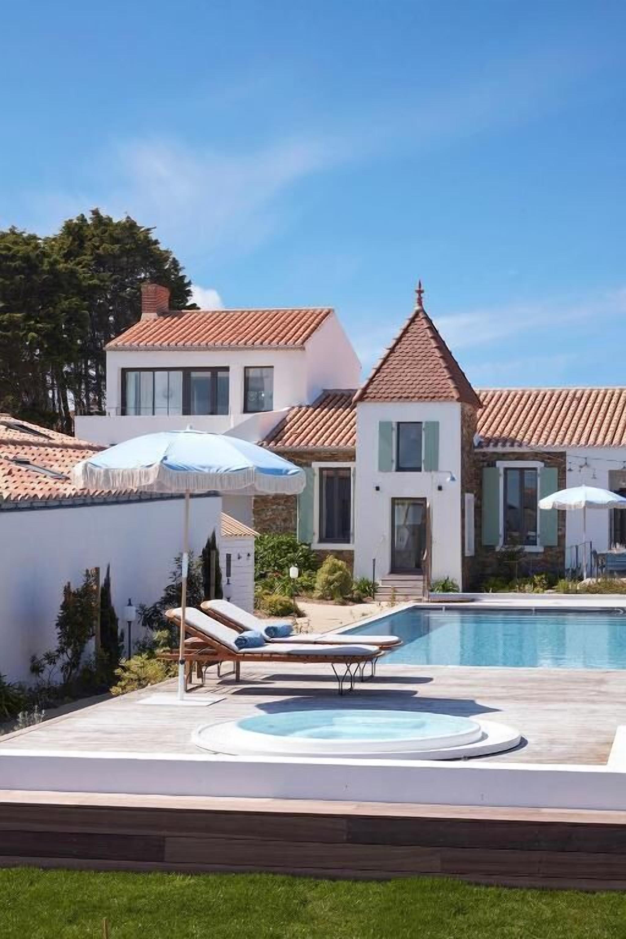 A swimming pool with lounge chairs and umbrellas, surrounded by a white building with a tiled roof.