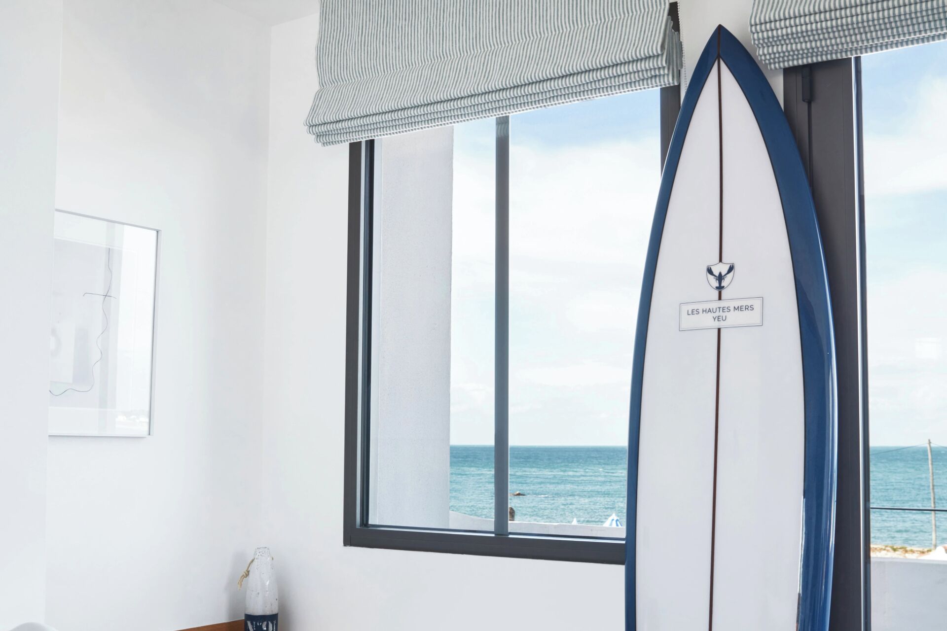 A room with a large window overlooking the sea, a surfboard with a logo, and a framed picture on the wall.