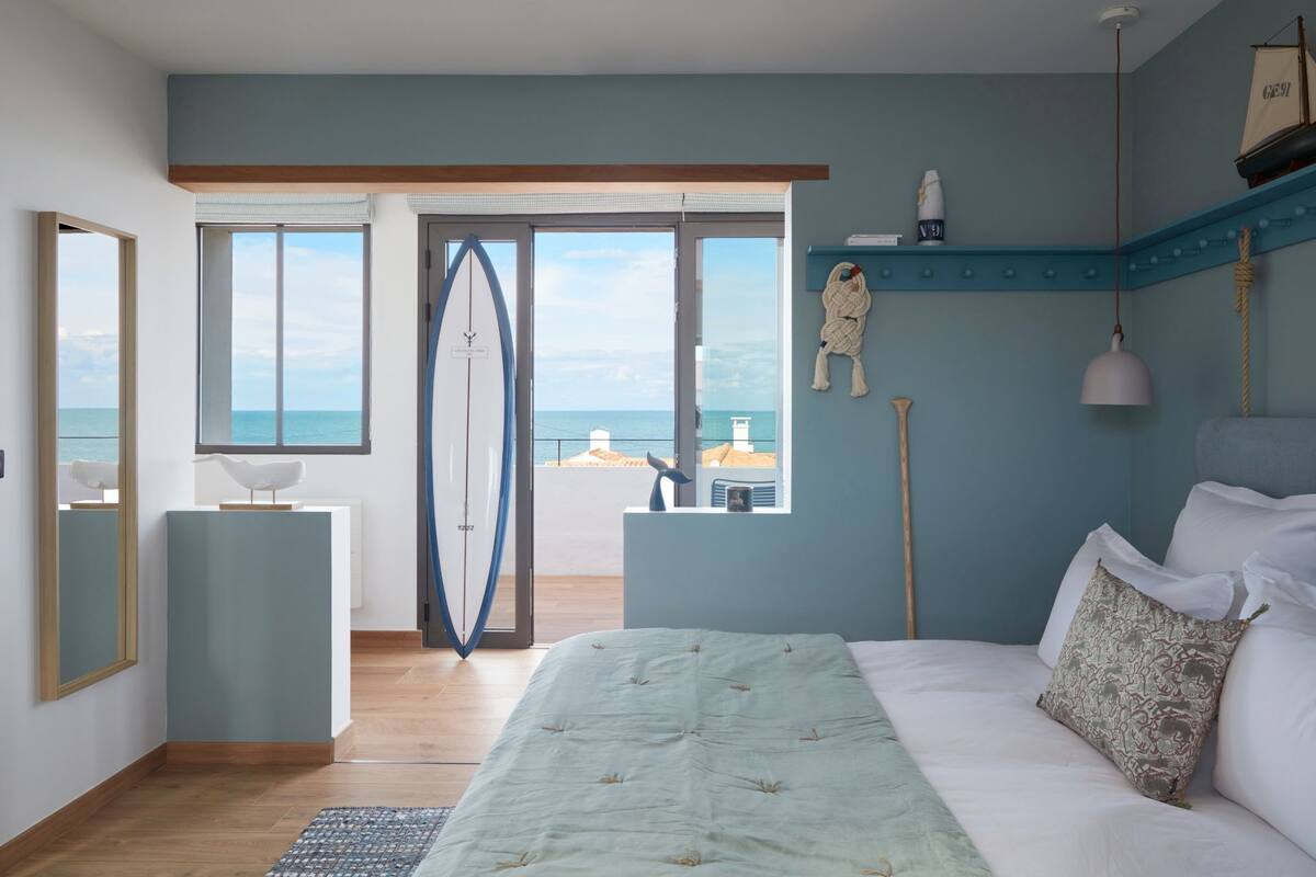 A bedroom with a bed, a window with a view of the sea, and a surfboard leaning against the wall.