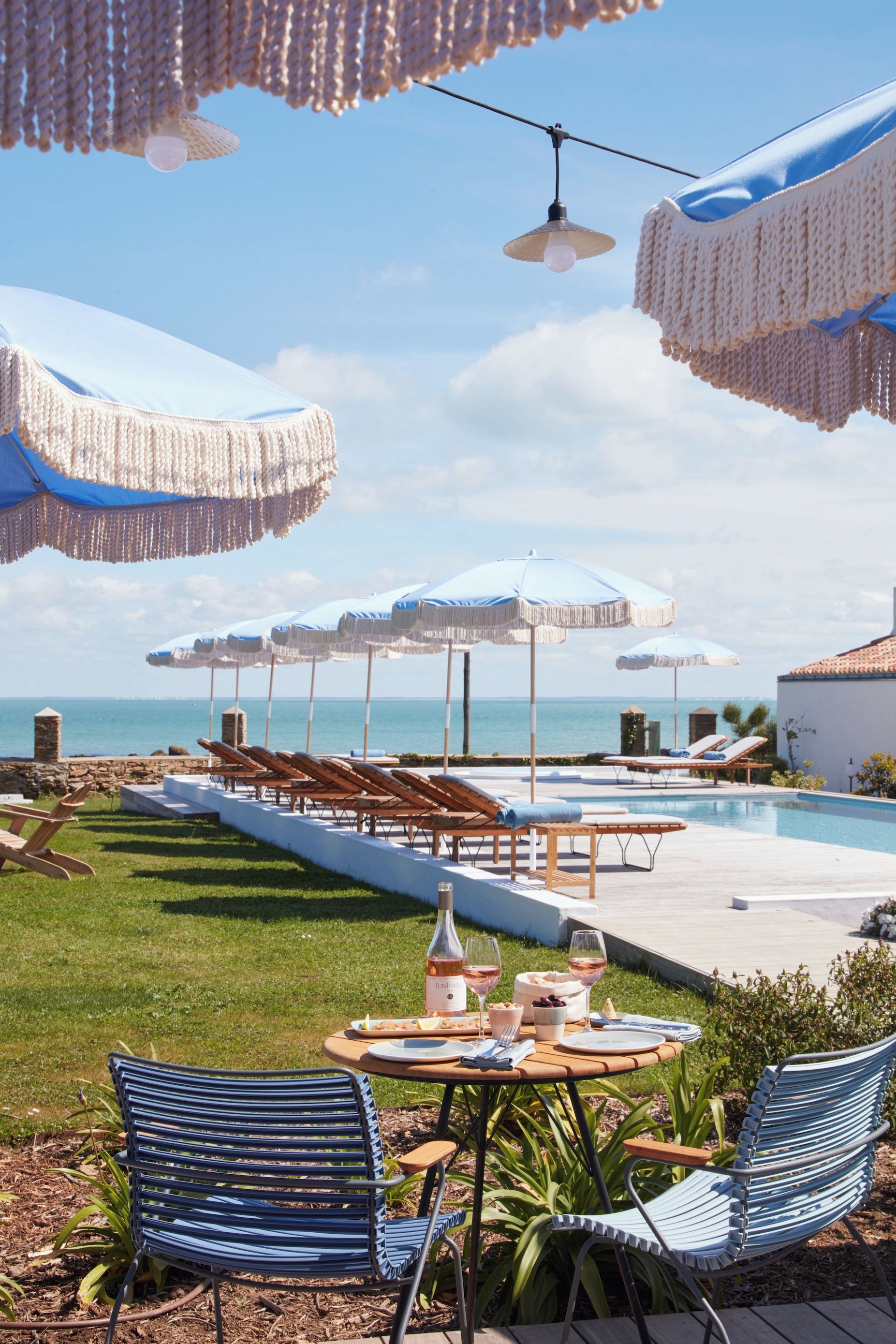 Zona de piscina con sillas reclinables, sombrillas y una mesa de comedor con una botella de vino.