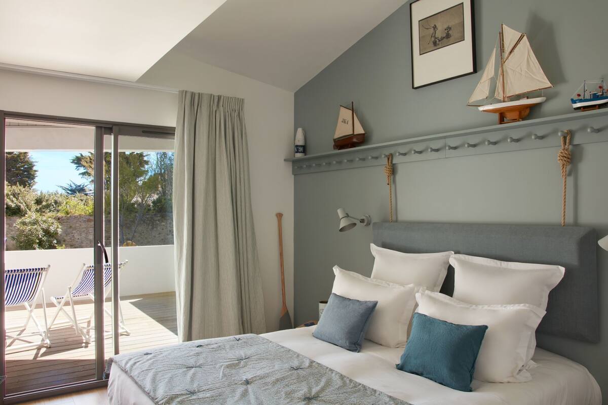 A bedroom with a bed, pillows, a headboard, a window with curtains, and a balcony with chairs.