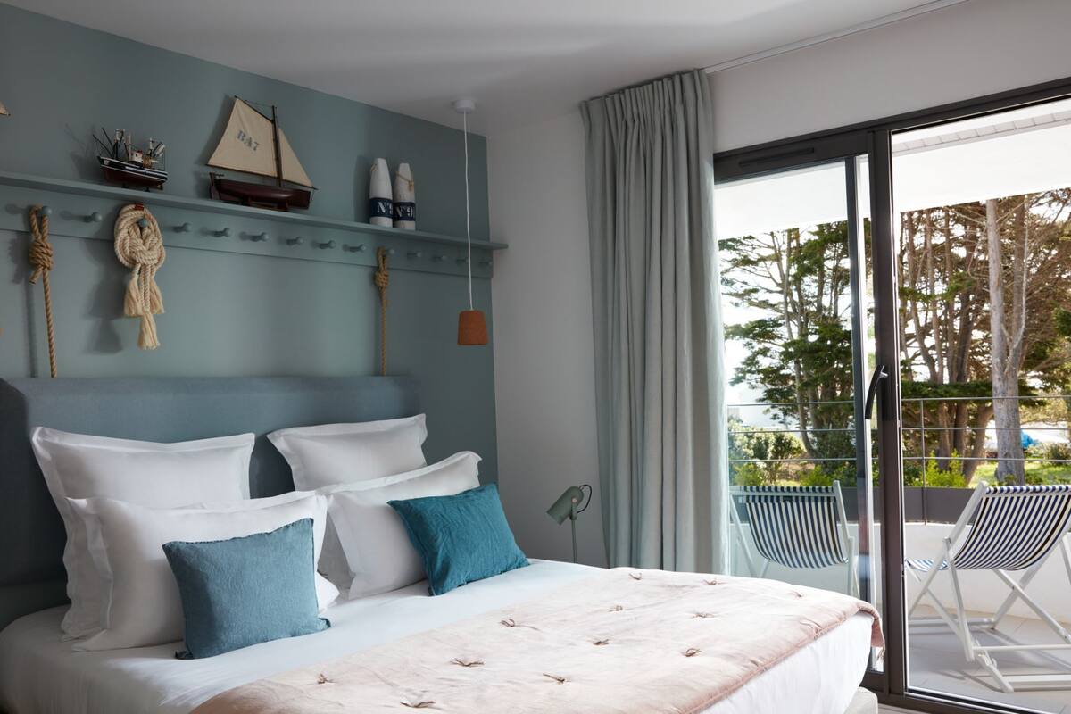 A bedroom with a bed, a headboard, a window with curtains, and a balcony with chairs.