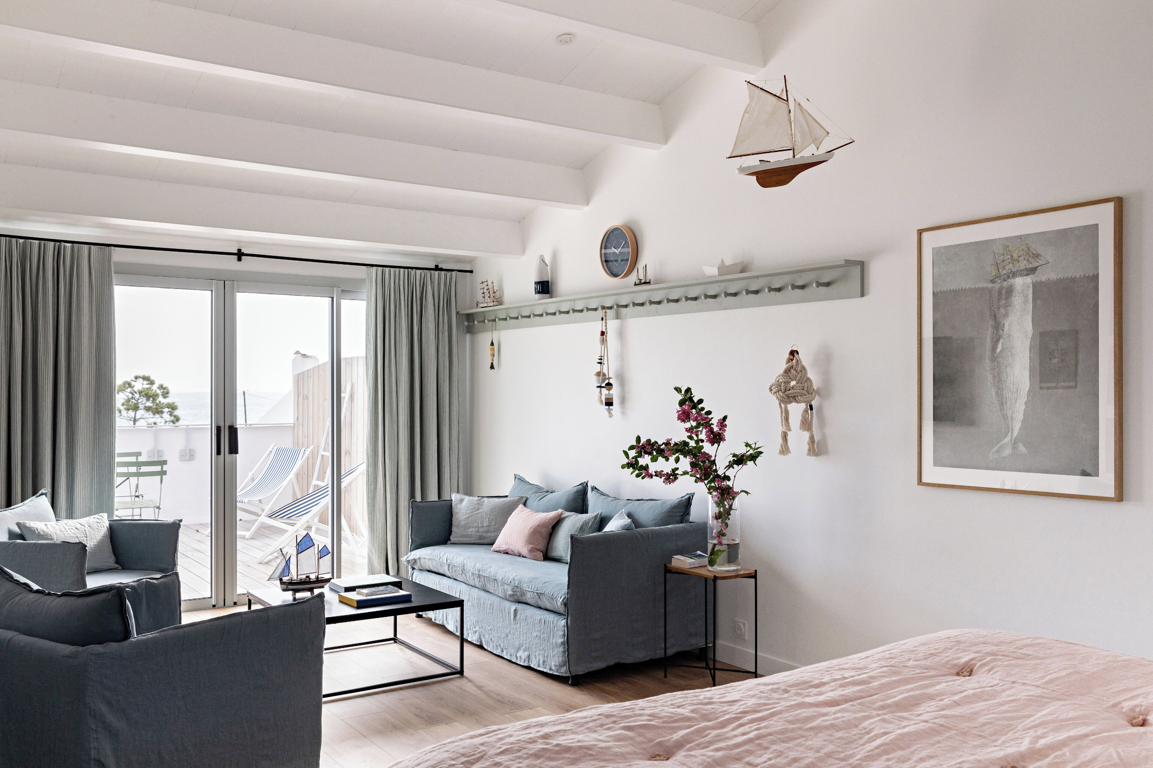 A modern living room with a sofa, armchairs, a coffee table, and a bed. There is a balcony with a view, a clock on the wall, and a framed picture.
